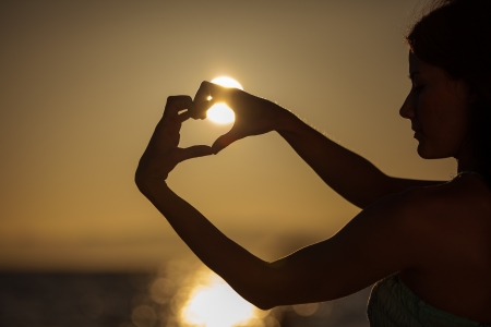 Silhouette of a young woman creating the shape of a heart with her hands with the sun nearly insideの写真素材
