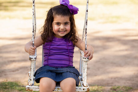 Cute little Latin girl having fun on a swing at a park and smiling