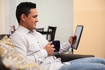 Relaxed young man having a cup of coffee and reading the news on a tablet computer at homeの写真素材
