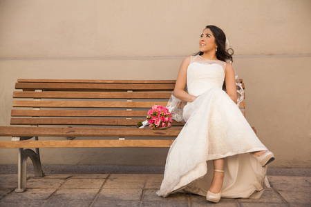 Cute Hispanic bride sitting on a bench outside and looking up towards copy spaceの写真素材