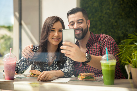 Attractive young Hispanic couple taking a selfie with a smartphone while eating lunch