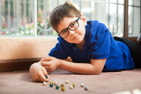 Portrait of a little boy with glasses lying on the carpet floor and playing with some marbles at homeの写真素材