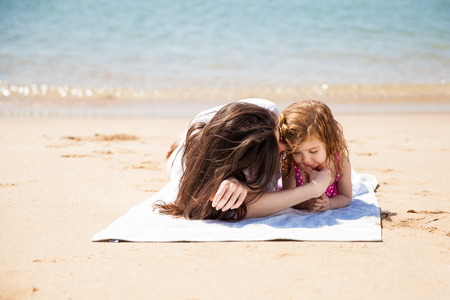 Cute little girl and her mother relaxing and lying on a towel on the sand on a sunny dayの写真素材