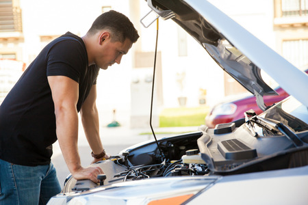 Profile view of a handsome young Hispanic man looking at a car with its hood open, trying to fix itの写真素材