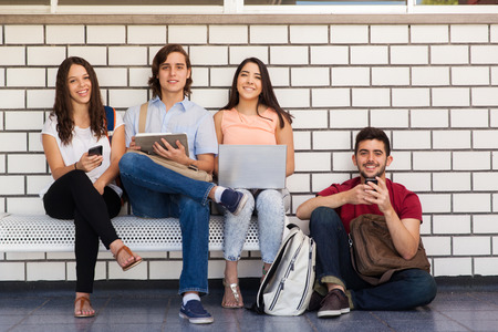 Full length portrait of a group of four students sitting on a school hallway and using all kinds of technologyの写真素材