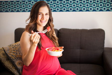 Goodlooking pregnant woman eating healthy food in her living room and smiling