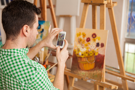 Young male artist taking a photo of his artwork with a smartphone to share it on a social network and sell it onlineの写真素材