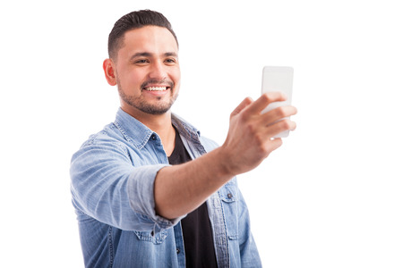 Handsome young Hispanic man taking a selfie with his smartphone against a white background