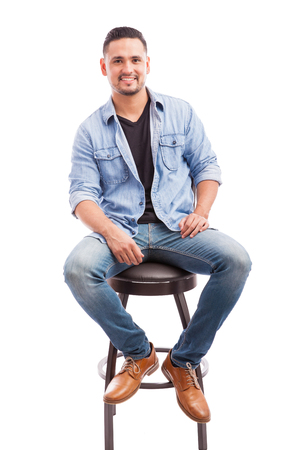 Attractive young man dressed casually sitting on a chair and smiling against a white backgroundの写真素材