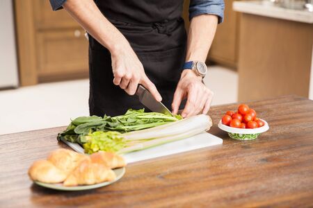 Closeup of a young man wearing an apron and chopping some vegetables to make a salad at home