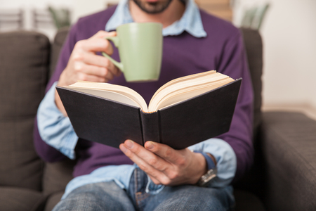 Closeup of a young man reading a book while drinking coffee from a mug at homeの写真素材