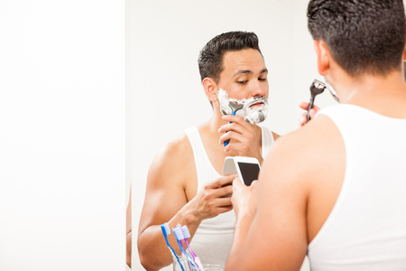 Portrait of a busy and distracted young man multitasking and shaving his beard while texting on his smartphoneの写真素材