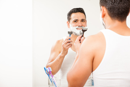 Handsome young man using a razor to shave his beard in the bathroom and smilingの写真素材