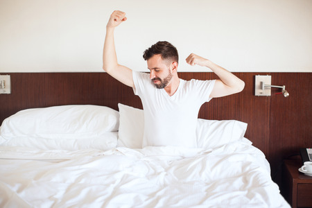 Handsome young man waking up and stretching on his bed right after waking up in the morningの写真素材