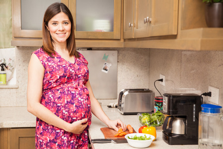 Gorgeous young Hispanic pregnant woman making a healthy salad in a kitchen and smilingの写真素材