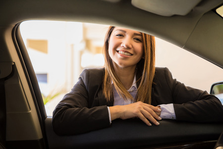 Gorgeous young female driver in a suit leaning on a car door and ready to drive you to your next destinationの写真素材