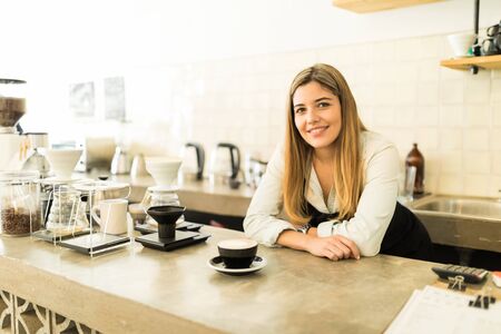Portrait of a gorgeous young Hispanic barista leaning on the counter in front of a cup of cappuccino in a coffee shopの写真素材