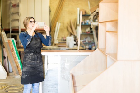 Pretty female carpenter using a smartphone to take a picture of some furniture she just made in her workshopの写真素材