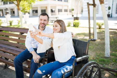 Happy young woman sitting on a wheelchair and taking a selfie with her smartphone of herself with a guy she is datingの写真素材