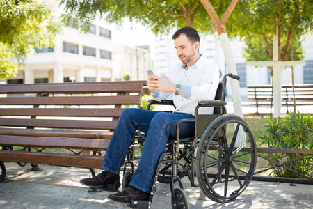 Full length view of a young man in a wheelchair using his smartphone to check on social media at a parkの写真素材