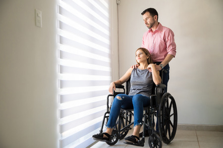 Pretty young disabled woman sitting on a wheelchair and holding hands with her partner at homeの写真素材