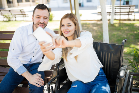 Beautiful Caucasian woman in a wheelchair taking a selfie with her date while they both sit in a parkの写真素材