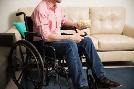 Closeup of a man on a wheelchair watching tv and eating popcorn at home to pass the timeの写真素材