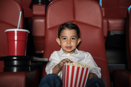 Portrait of a good looking Hispanic boy sitting in a movie theater and eating a lot of popcornの写真素材