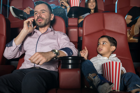 Little boy looking disappointed with his dad and asking him to be quiet while he talks on the phone at the movie theaterの写真素材