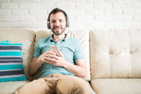 Hispanic young man relaxing at home listening to music in his phone with headphones and making eye contactの写真素材
