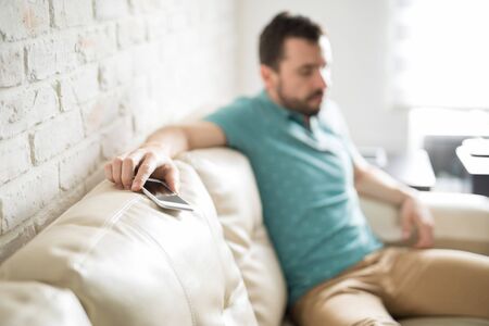 Hispanic young man sitting on the couch with his phone on his hand looking very sad about a call he just receivedの写真素材