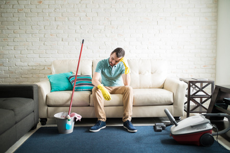 Latin man sitting in the living room tired of a long afternoon cleaning his house all by himselfの写真素材