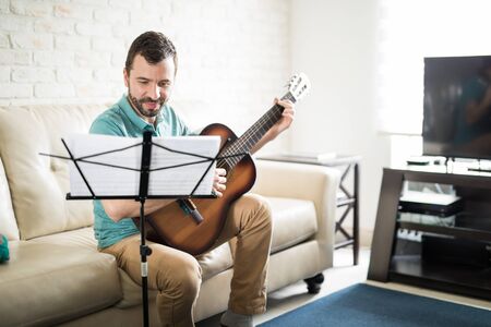 Attractive young Latin man playing the guitar and reading some music sheets in the living room