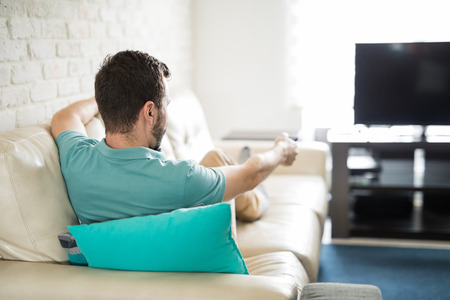 Man relaxing at home sitting in his cozy sofa holding a remote control in his hand and deciding what to watch on tvの写真素材