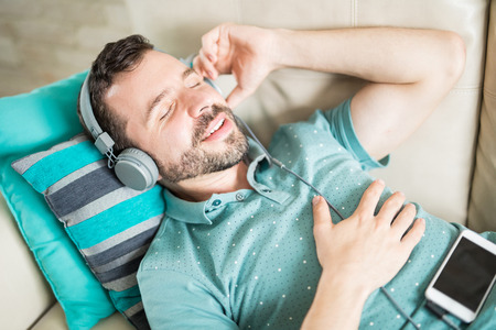 Latin man with his eyes closed enjoying great music in his phone and relaxing lying down on the couchの写真素材