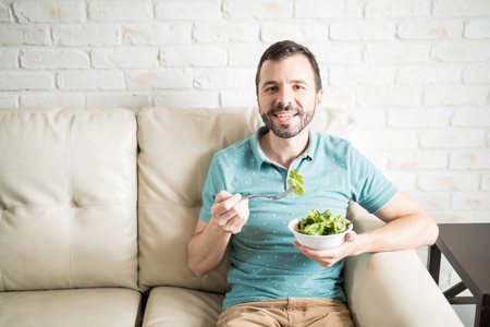 Healthy man eating a fresh and green salad for lunch alone at his apartment in the living roomの写真素材