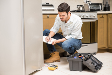 Portrait of a handsome male technician doing a work report on a broken fridge in a home kitchenの写真素材