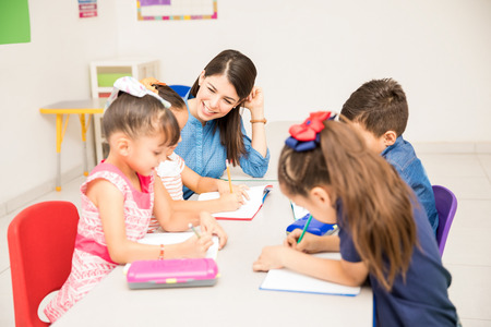 Happy kindergarten female teacher helping her students with some writing exercise in the classroomの写真素材