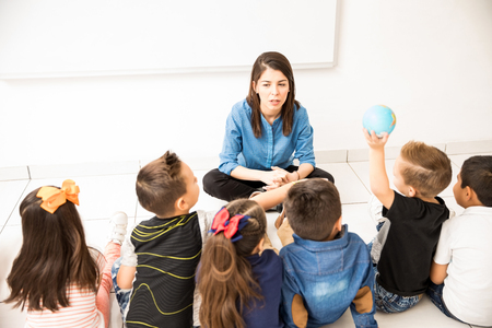 Attractive young woman teaching her preschool students while they all sit on the classroom floorの写真素材
