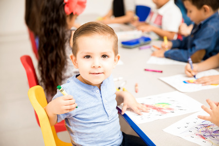 Handsome kindergarten male student doing some coloring with crayons in the classroom and having a good timeの写真素材
