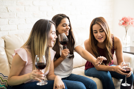 Friends drinking wine together at home while looking surprised at their friend's engagement ring
