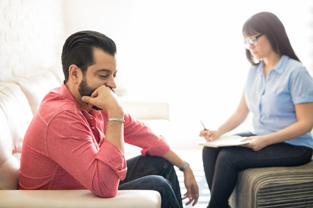 Young hispanic man with depression sitting on couch with female psychotherapist making notesの写真素材