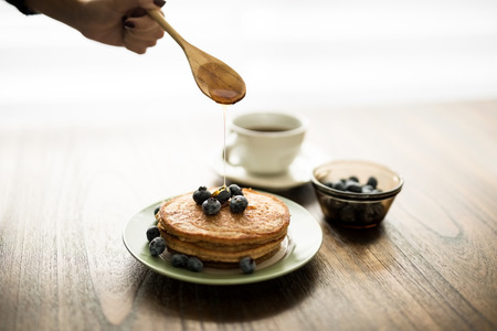 Woman putting honey over delicious pancakes with blueberries