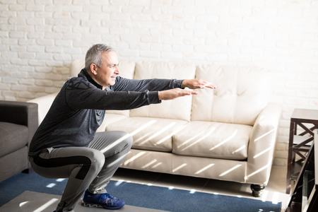 Senior man doing squats and exercising in living room at homeの写真素材