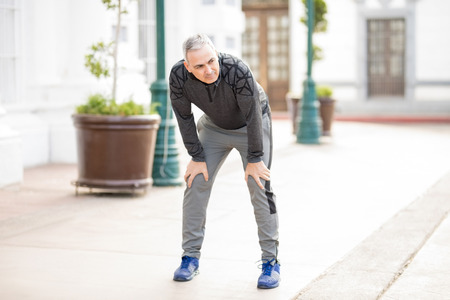 Tired middle aged hispanic man stopping with hands on knees to grab some air after working out in the cityの写真素材