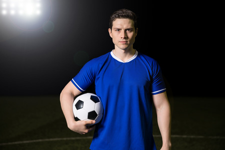 Portrait of handsome young soccer player making an eye contact while standing in blue uniform holding a ball in hand on soccer fieldの写真素材