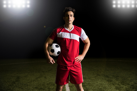 Soccer player wearing red uniform standing on football pitch under lights holding a ballの写真素材