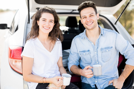 Beautiful young couple sitting with coffee in car trunk making an eye contact, break on road trip.の写真素材