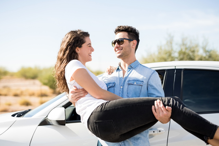 Handsome young man carrying his attractive girlfriend and smiling while standing next to a modern carの写真素材