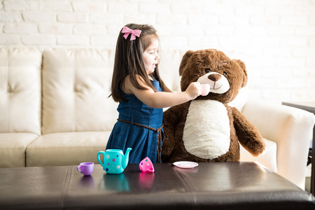Pretty little girl playing with her teddy bear at tea party using child's tea set. Young girl child happily feeding her teddy bear in living room.の写真素材
